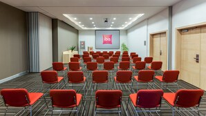 A conference room with rows of red chairs, a podium, a screen displaying 'ibis Hotels', and a ceiling with recessed lighting. - ibis Istanbul Esenyurt (Istanbul)