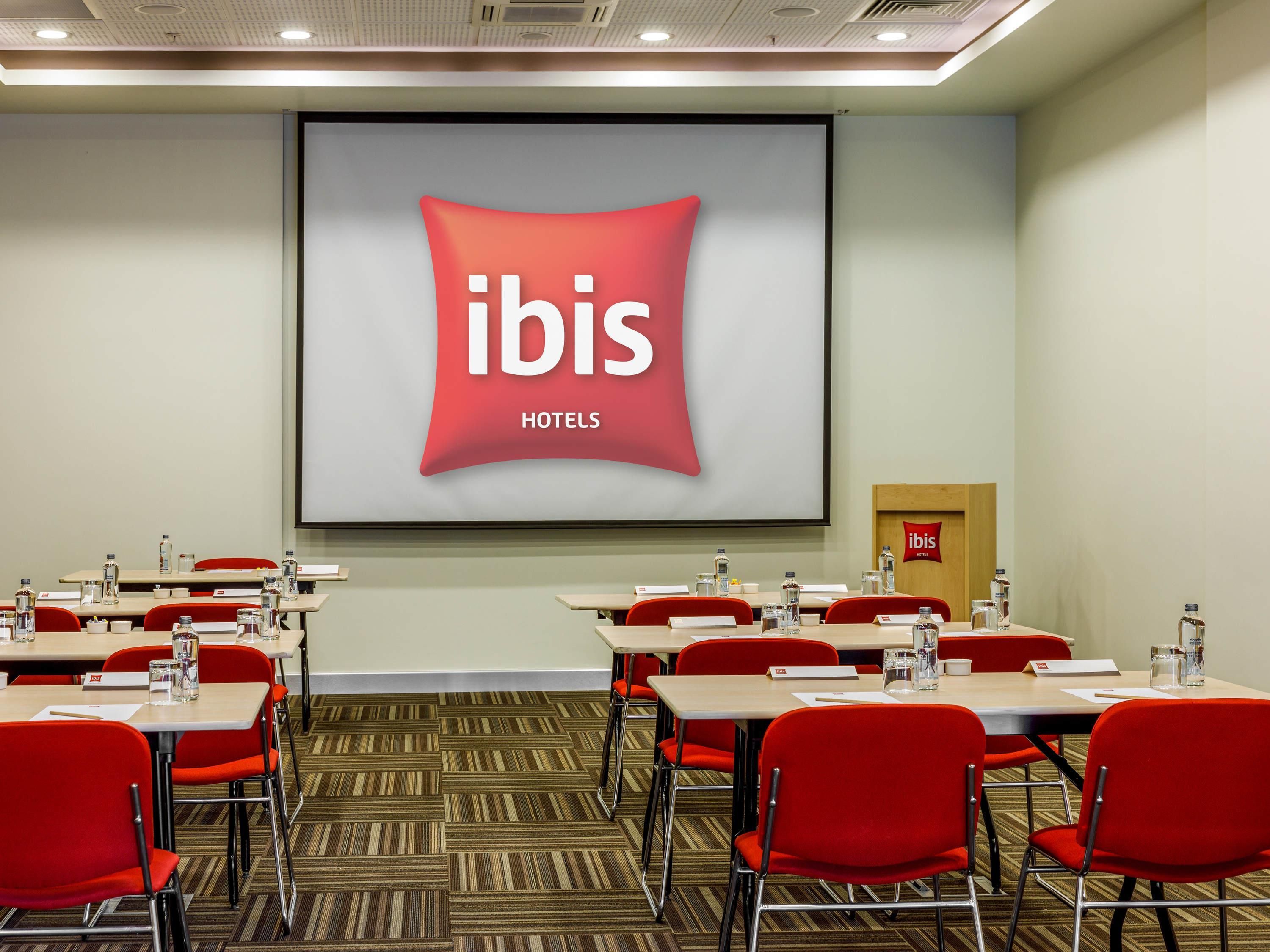 A conference room with a large screen displaying the ibis hotels logo, red chairs, and tables with water bottles.