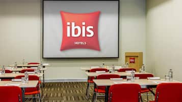 Un salón de conferencias con una pantalla grande mostrando el logotipo de ibis hotels, sillas rojas y mesas con botellas de agua.