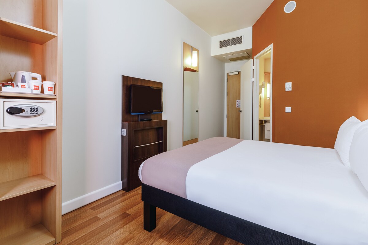A hotel room with a bed, a television, a minibar, and a wooden shelf with toiletries.