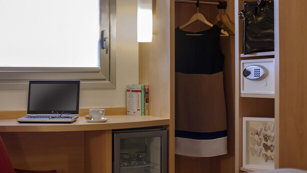 A hotel room with a laptop on a desk, a small refrigerator, a red chair, a wardrobe with a hanging dress, and a framed butterfly artwork. - ibis Istanbul Esenyurt (Istanbul)