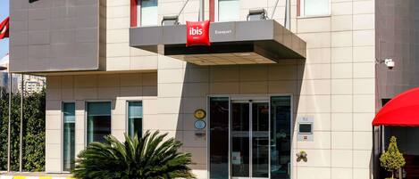 Una entrada de hotel moderna con un logotipo rojo llamativo, un vestíbulo cubierto y una palmera en primer plano.