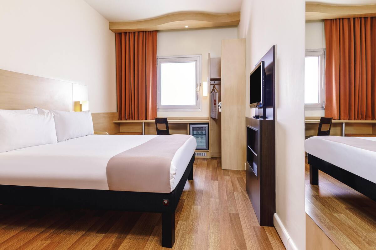 A hotel room with a bed, a TV, a desk, and a window with curtains.