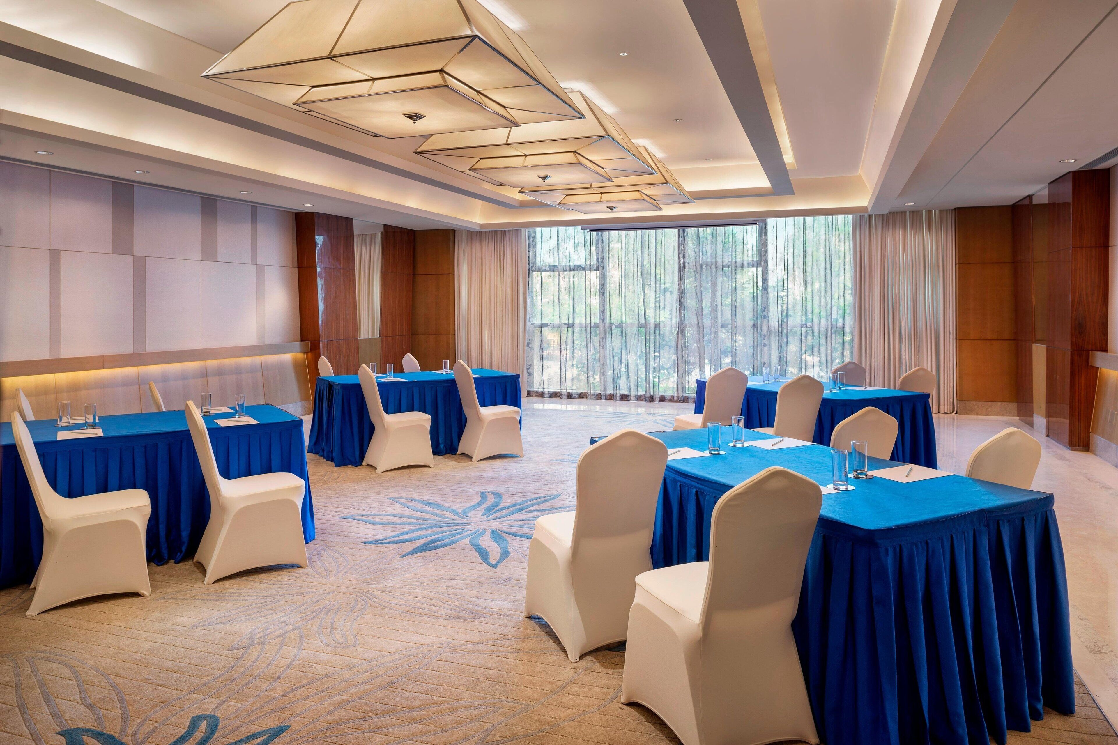 A conference room with multiple tables and chairs, large windows with curtains, and a patterned carpet.