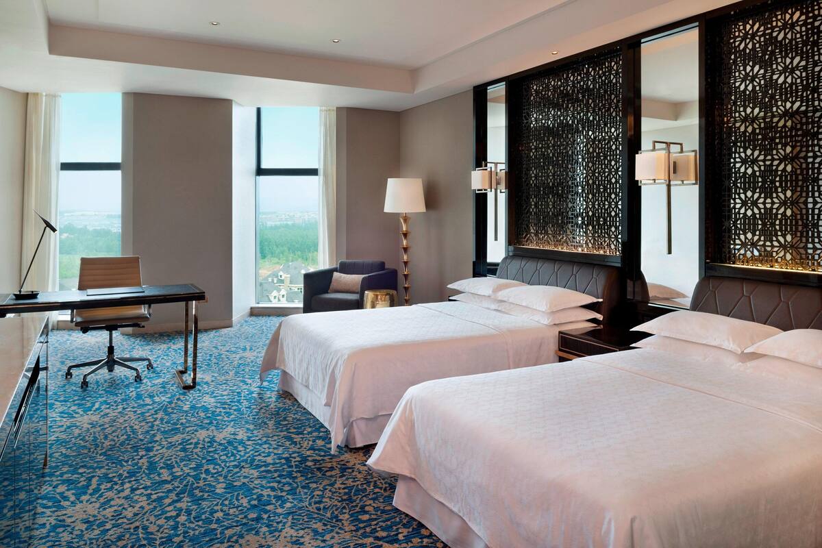 Een hotelkamer met twee bedden, een bureau, een stoel, een lamp en een groot raam met uitzicht op de stad.