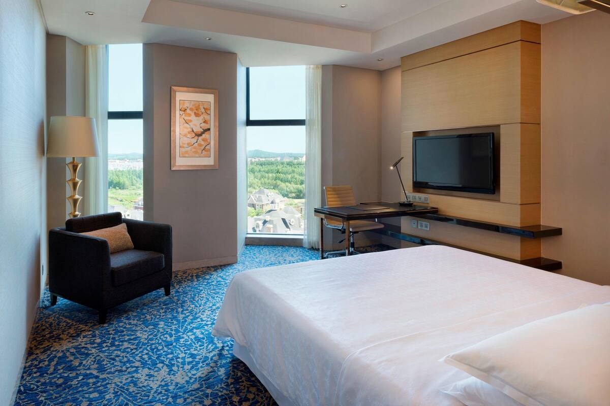 Een hotelkamer met een groot bed, een bureau met een flatscreen televisie, een stoel en uitzicht op de omgeving door grote ramen.