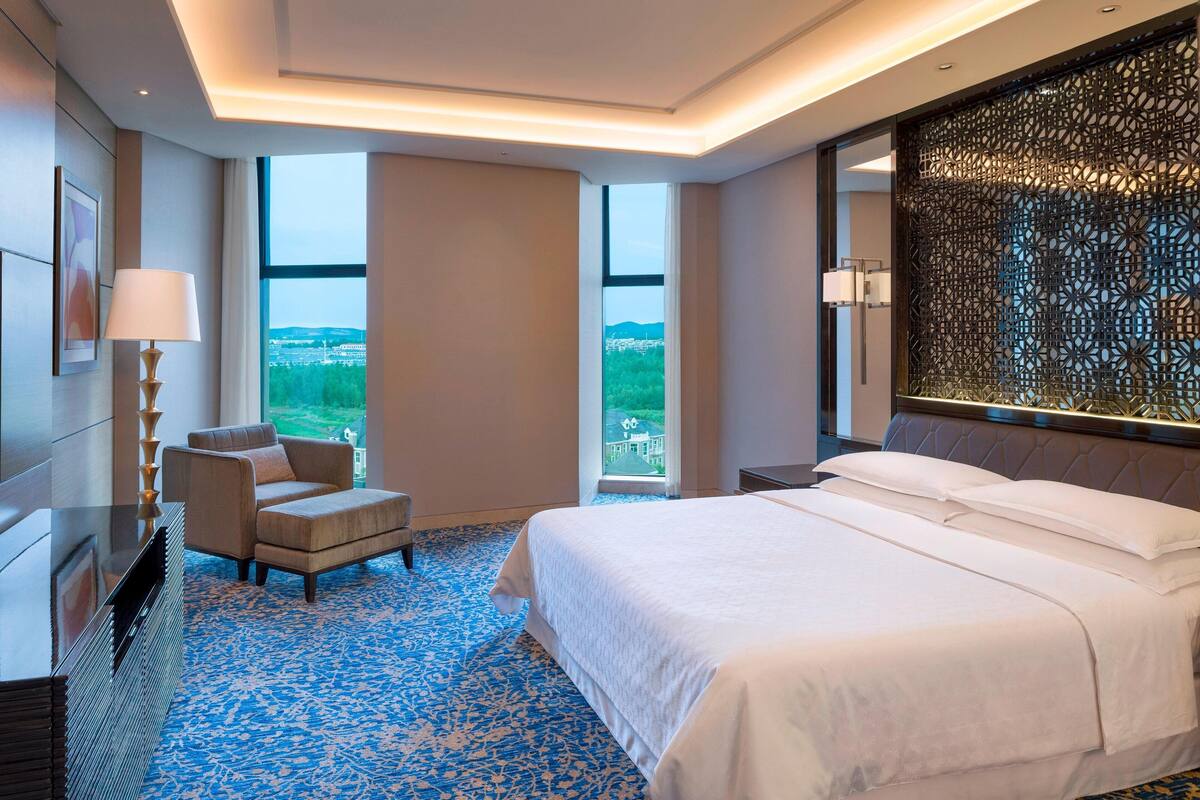 Een moderne hotelkamer met een groot bed, een zithoek met een stoel, een televisie en een uitzicht op een stadsbeeld door grote ramen.