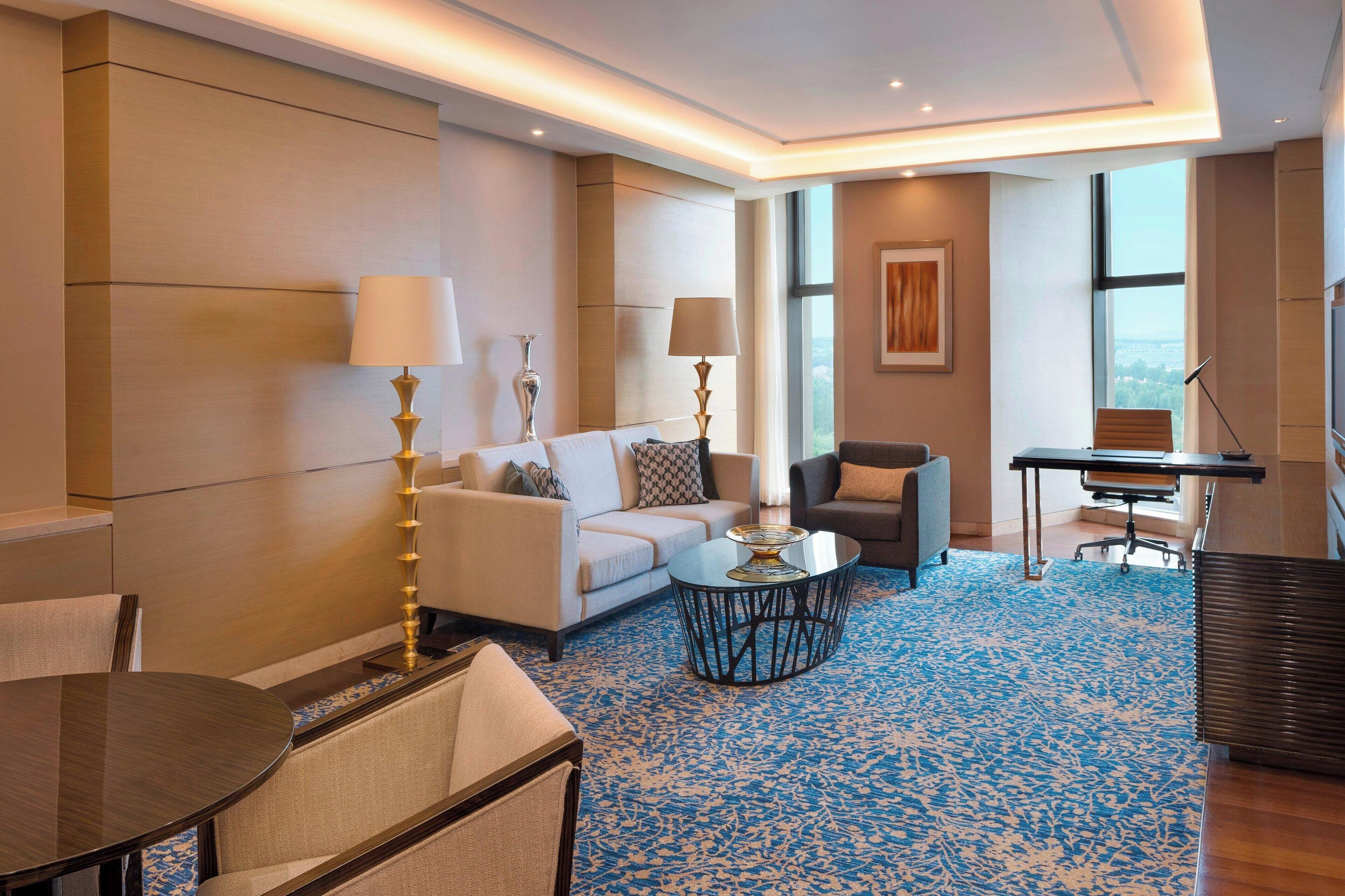 A modern hotel room with a sofa, armchair, and a small dining table. There is a large window providing a view of the city.