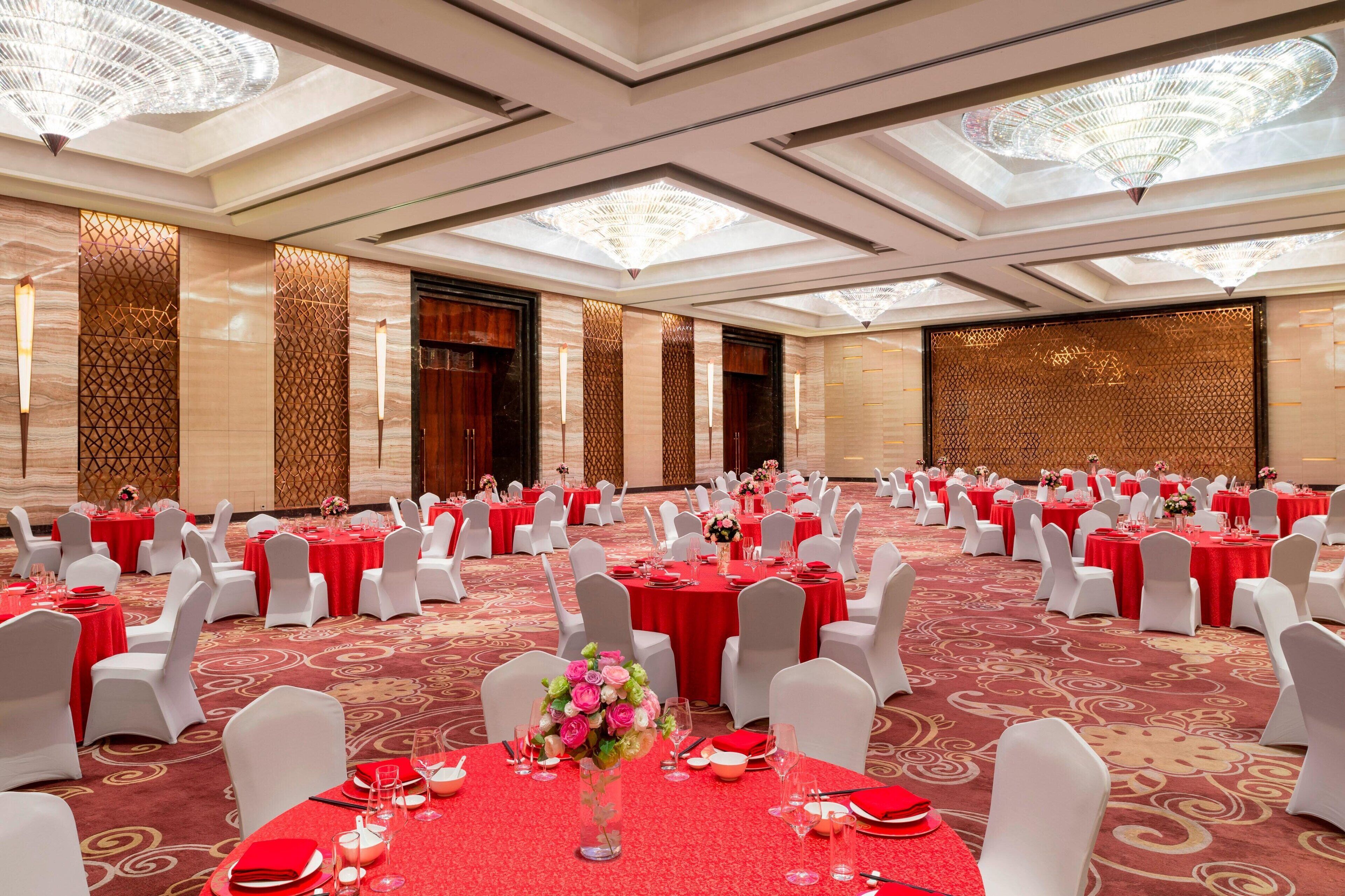 A banquet hall with round tables set for dining, white chairs, and red tablecloths.