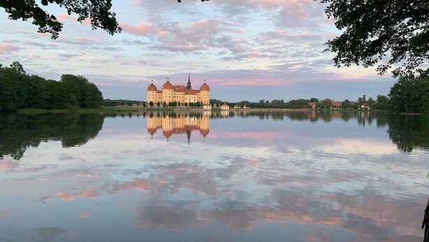 A large, historic building with a symmetrical facade and multiple towers, reflecting in a calm lake. - Pension Marlis (Moritzburg)