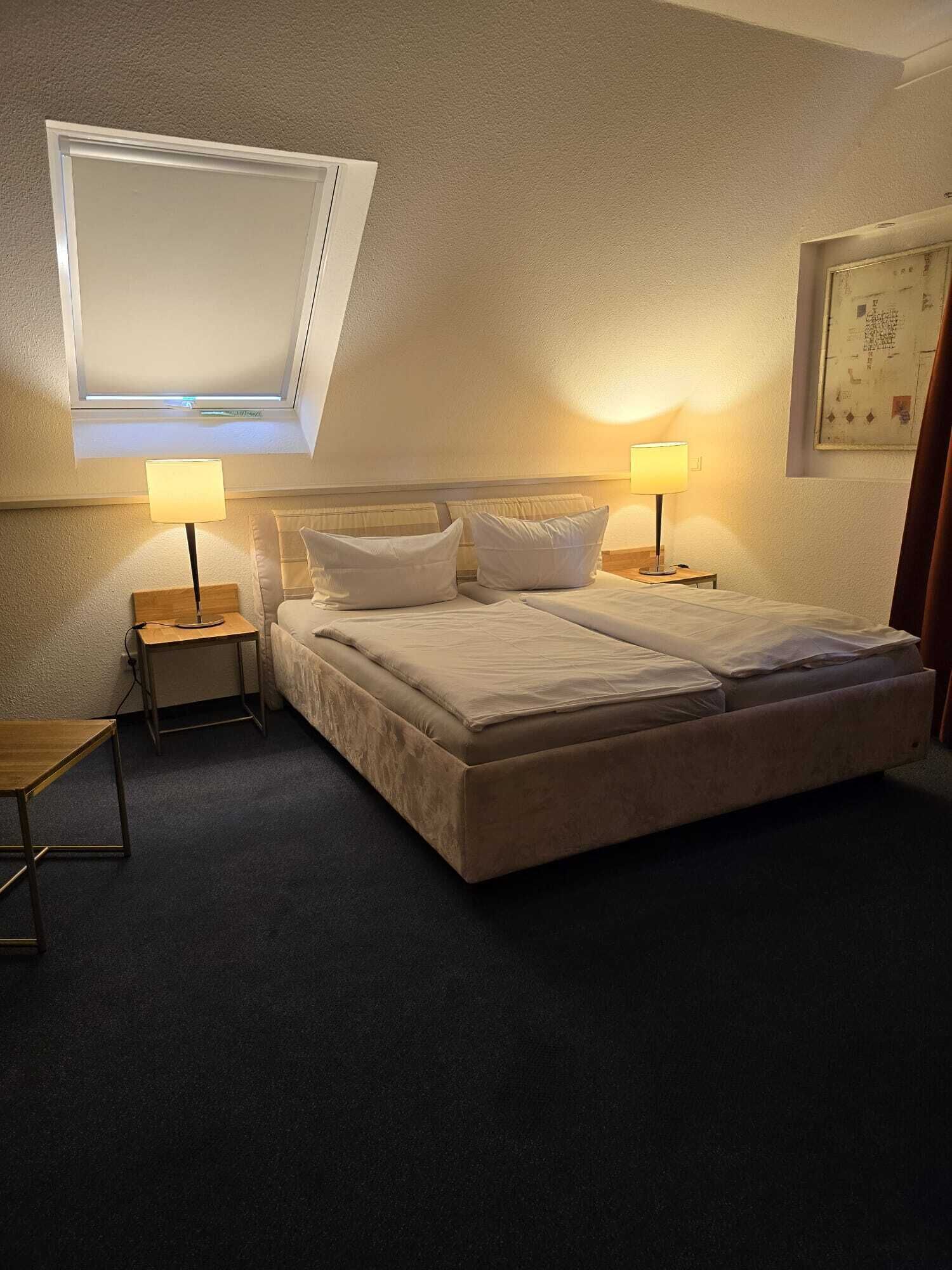 Habitación de hotel con una cama grande, dos mesitas de noche, un escritorio pequeño y un tragaluz sobre la cama.