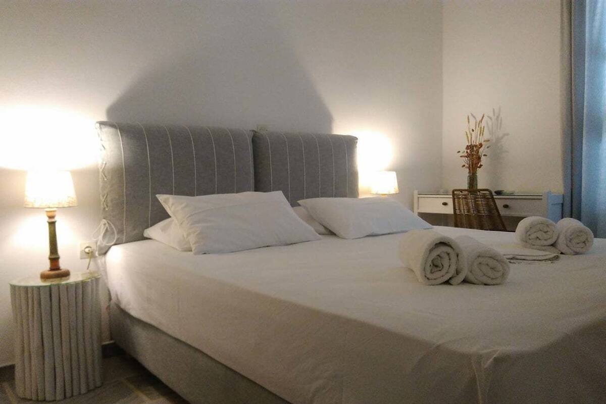 A neatly made bed with white linens and rolled towels, a bedside lamp, and a vase with dried plants on a nightstand.
