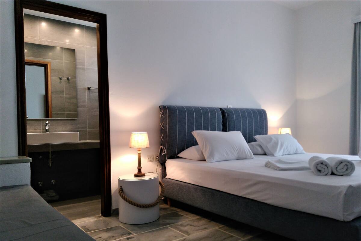A modern hotel room with a large bed, a bedside table with a lamp, a round bedside table with a rope handle, a large mirror, and a bathroom with a sink and mirror.