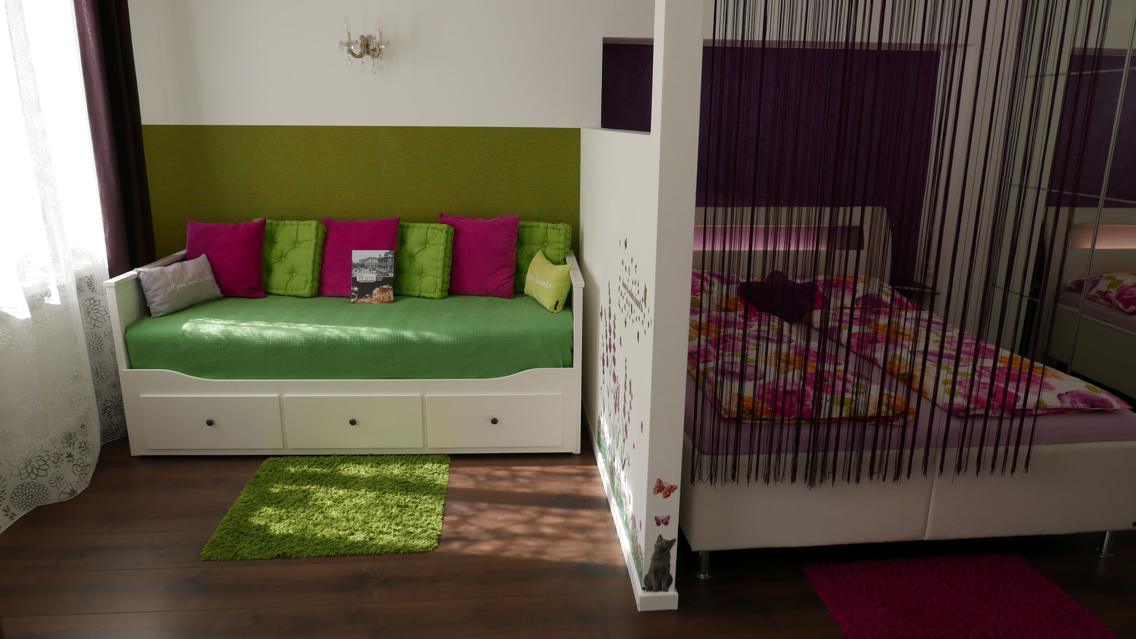 Un dormitorio con una cama verde, mesitas de noche blancas y un dosel morado.