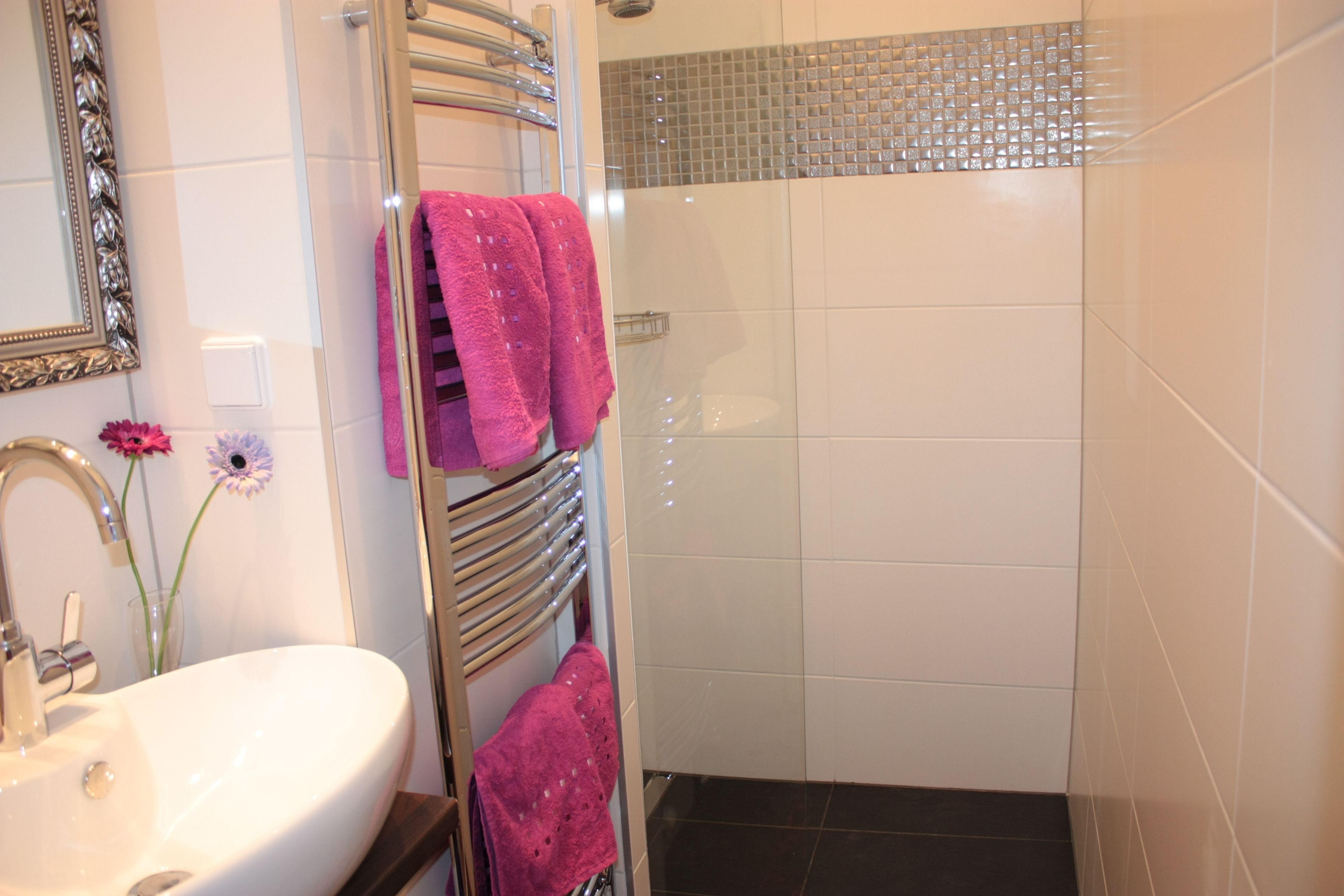 A bathroom with a white sink, a mirror, a glass vase with flowers, and a shower area with a glass door.