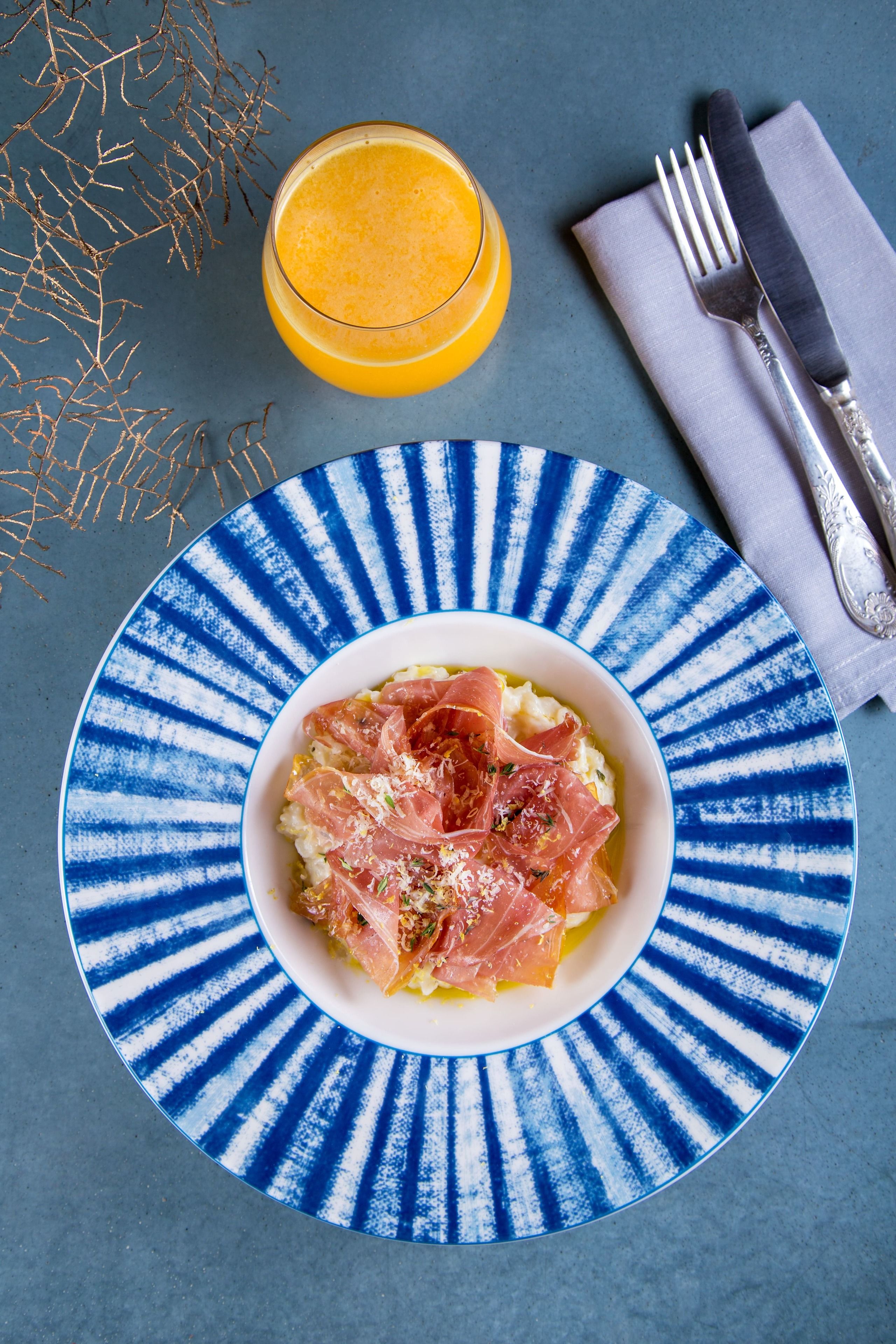 A plate of food with a blue and white patterned rim, a fork, and a knife on a napkin, with a glass of orange juice in the background.