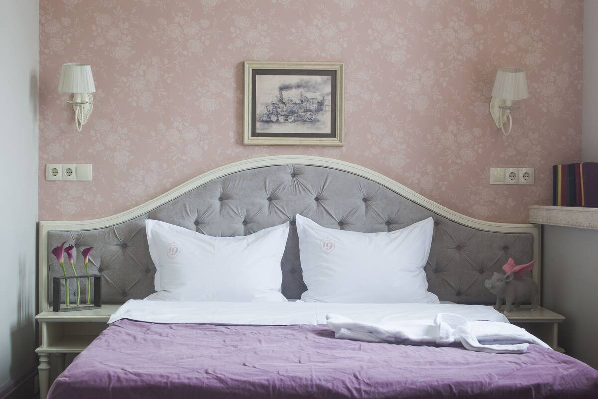 Una cama bien hecha con colcha morada, cabecera capitonada y dos mesitas de noche con lámparas y un cuadro enmarcado encima.