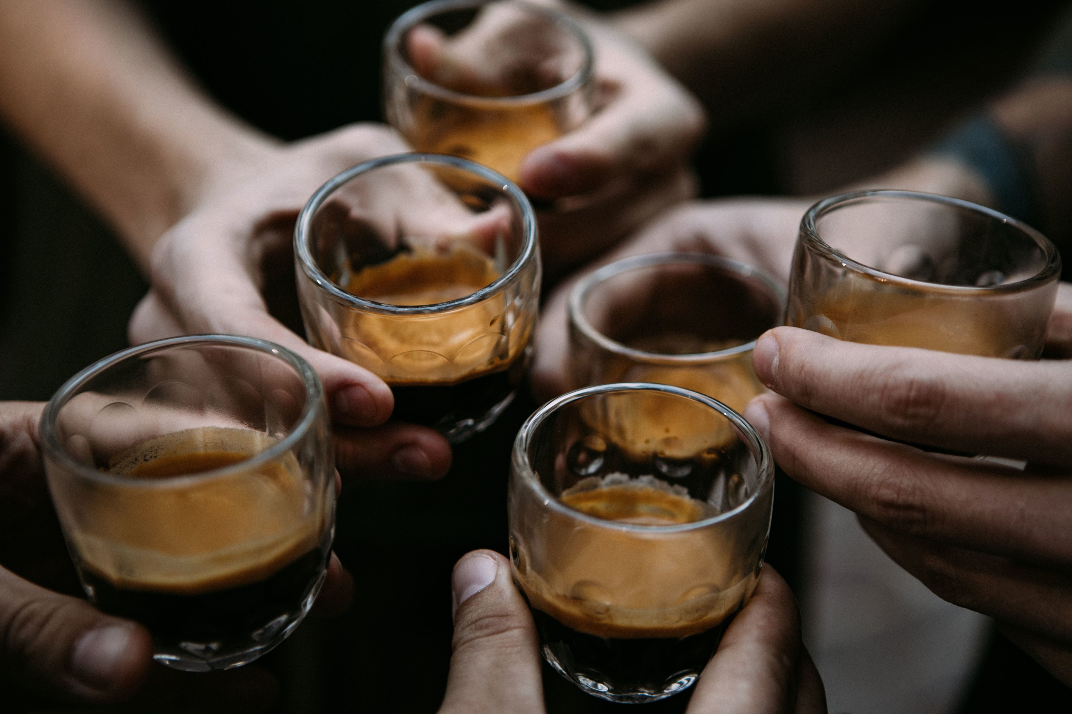 A group of hands holding shot glasses filled with a dark liquid, likely coffee or a similar beverage.