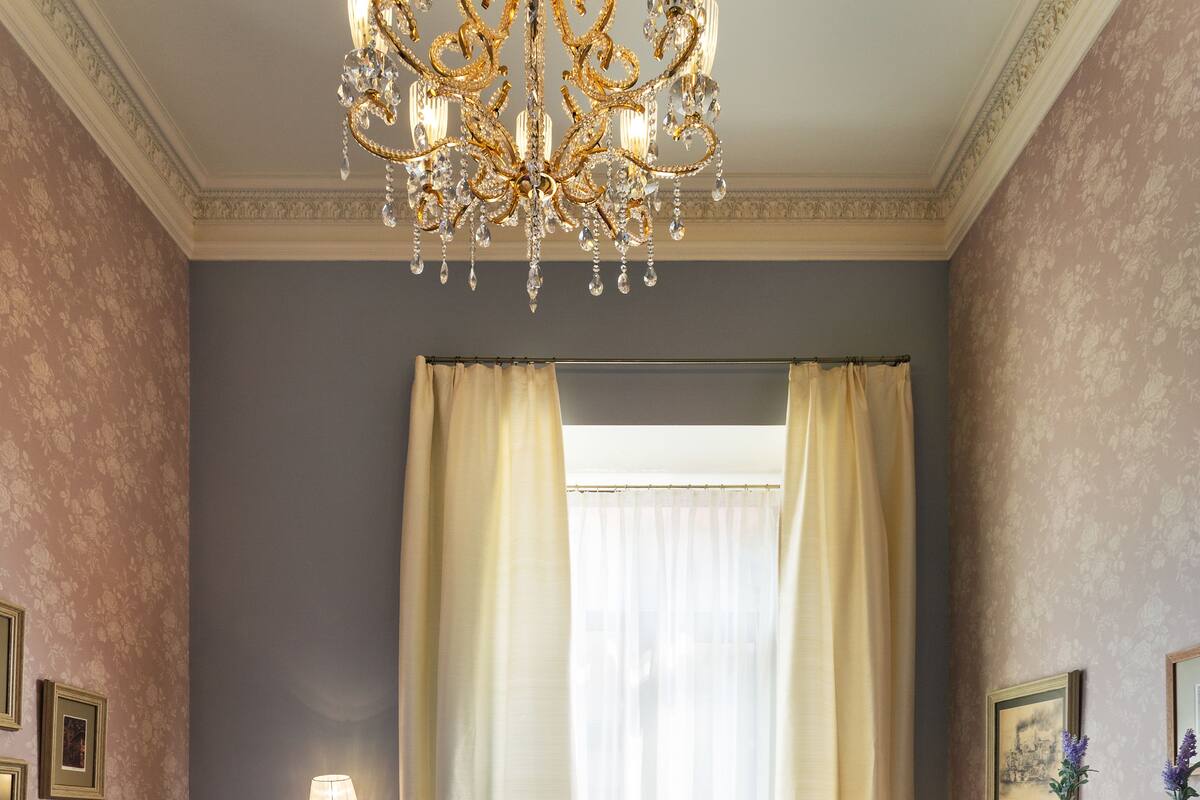Una habitación con una cama, una silla, una mesita pequeña con una jarrón y vasos, una estantería y una ventana con cortinas.