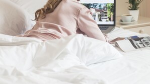 Una persona acostada en la cama con una laptop sobre una mesita de noche, una taza de café y una planta.