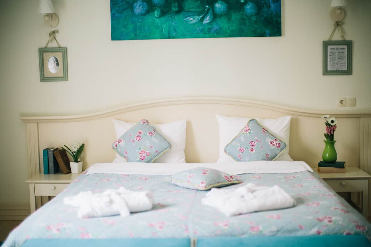 A neatly made bed with floral bedding, a vase with flowers, and a framed picture on the wall.