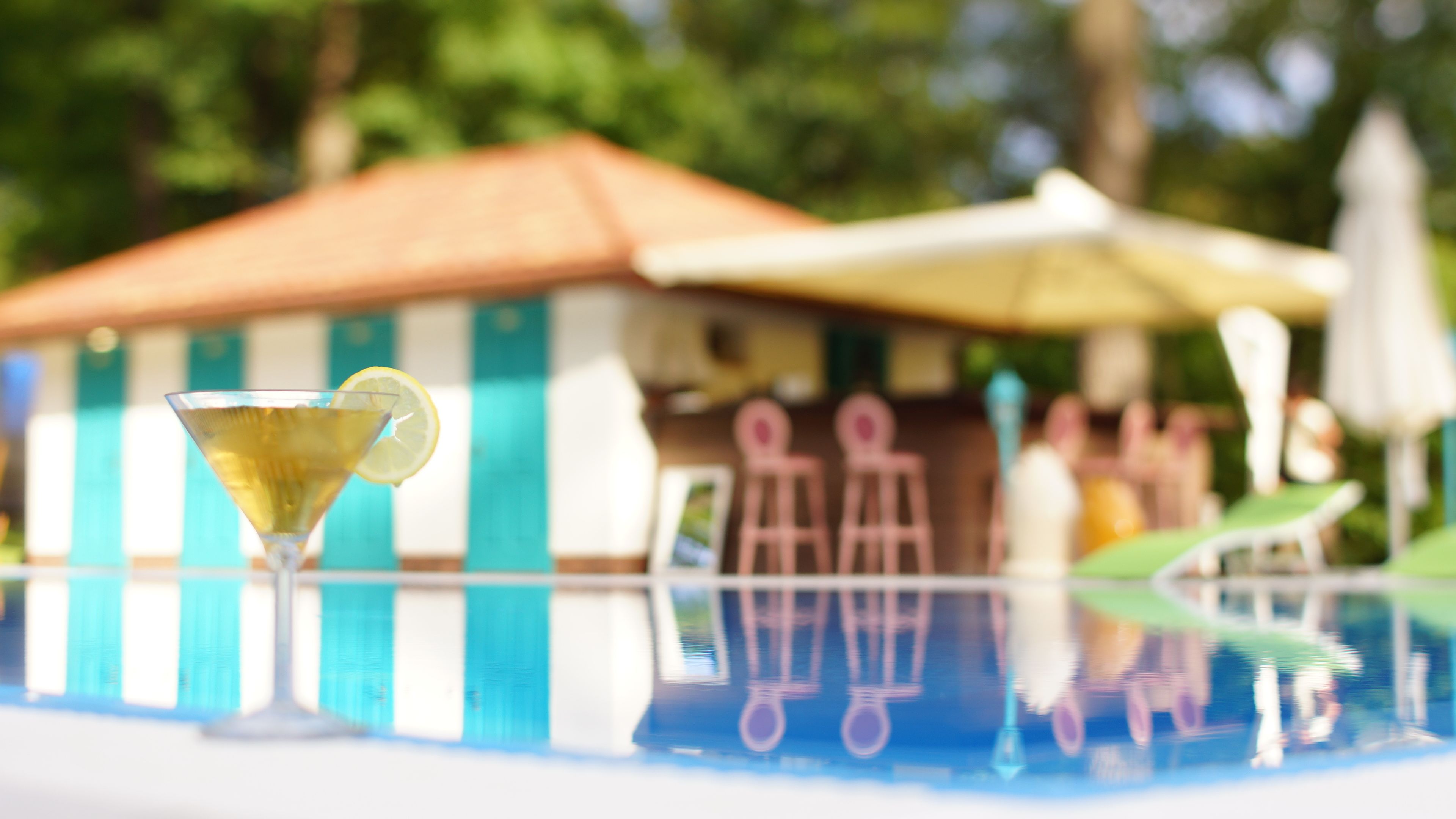 Un verre à cocktail, orné d’une tranche de citron, est posé sur une table près d’un espace aménagé autour d’une piscine, avec un bâtiment à rayures et des parasols.