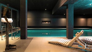A modern indoor pool area with a clear pool, lounge chairs, and a fire feature on the wall. - Hotel Galery69 (Stawiguda)