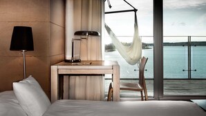 A hotel room with a bed, a desk, a lamp, a chair, and a view of a body of water through large windows. - Hotel Galery69 (Stawiguda)