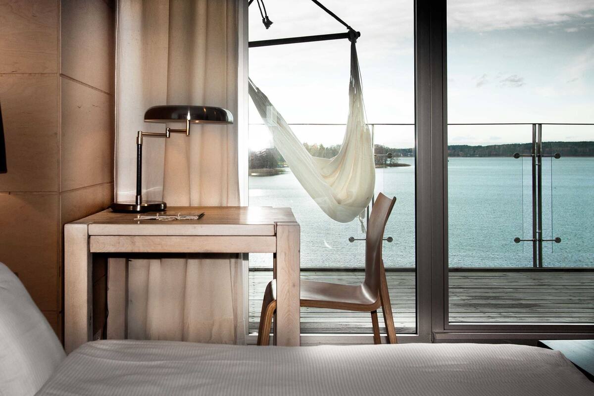 Habitación de hotel con cama, escritorio, lámpara, silla y vista a un cuerpo de agua a través de amplias ventanas.