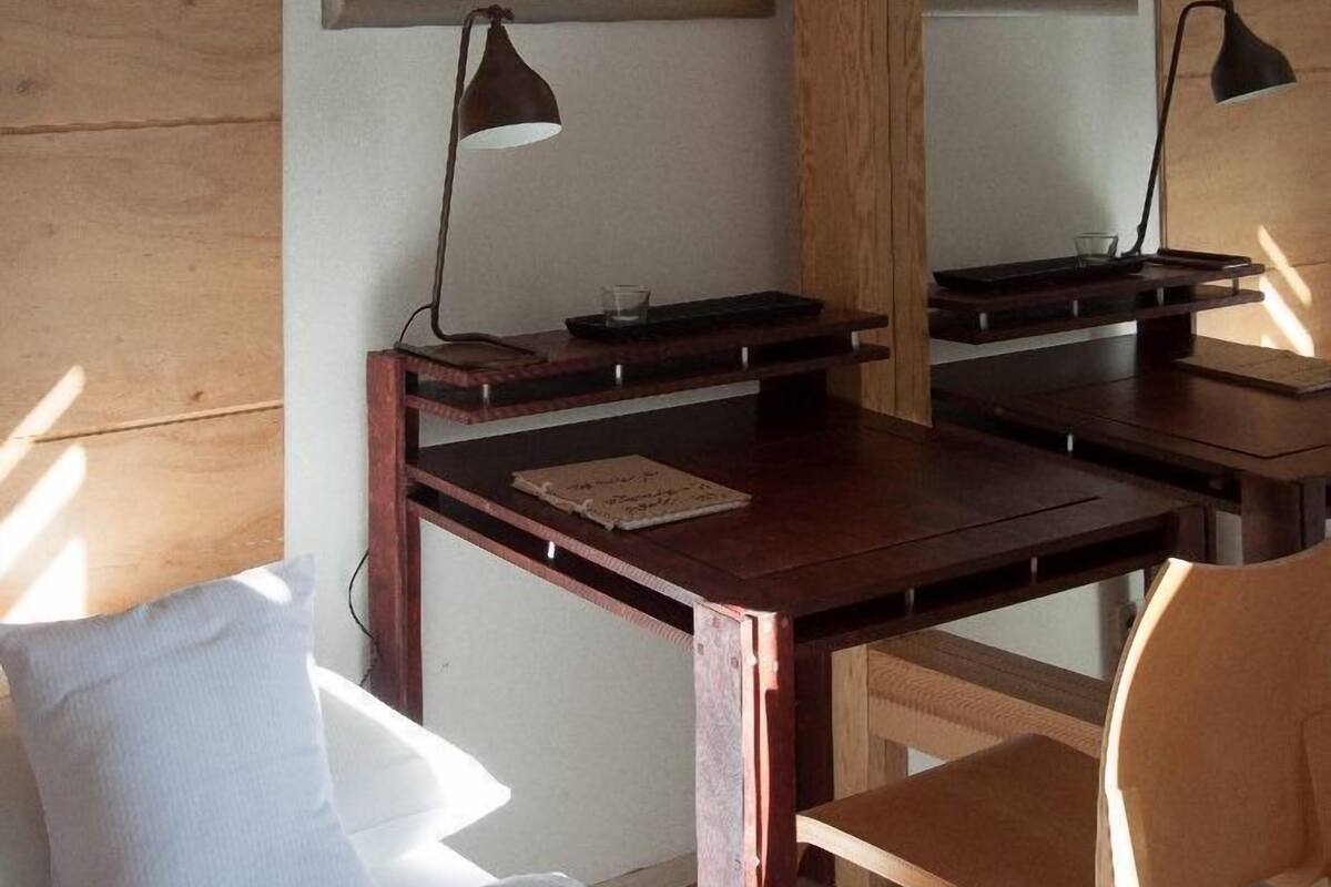 Una habitación con un escritorio de madera, dos sillas, una cama con sábanas blancas y dos cuadros en la pared.