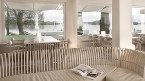 A restaurant with a view of a lake, featuring white furniture and a large window. - Hotel Galery69 (Stawiguda)