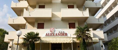 A multi-story hotel named "ALEXANDER" with a central entrance, multiple balconies, and a staircase leading to the entrance.