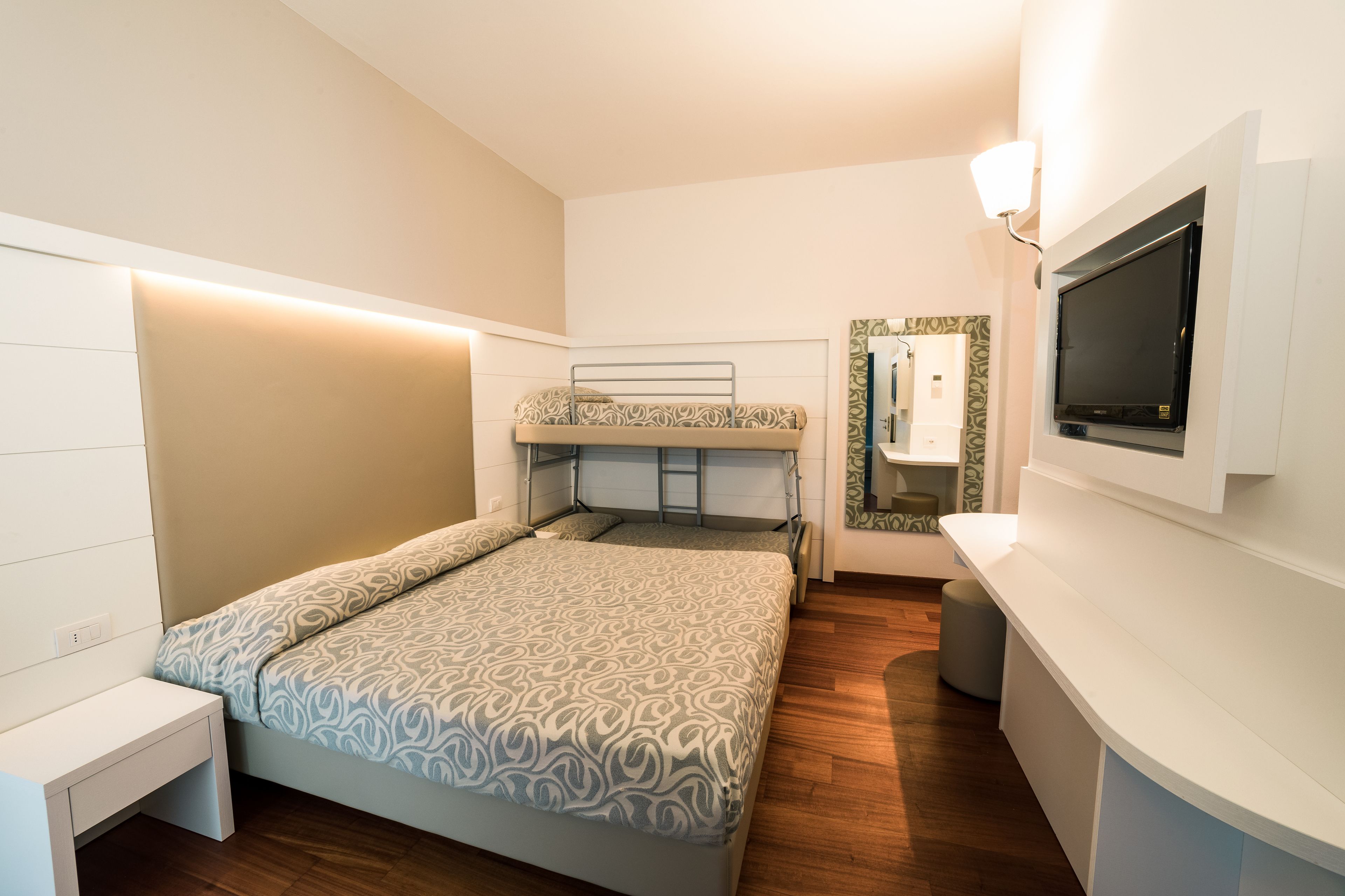 Una habitación pequeña con una cama litera, una mesita de noche, un televisor montado en la pared y un taburete.