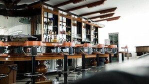 Un’area bar con diversi sgabelli e un bancone, travi in legno a soffitto, e scaffali con bottiglie e bicchieri.
