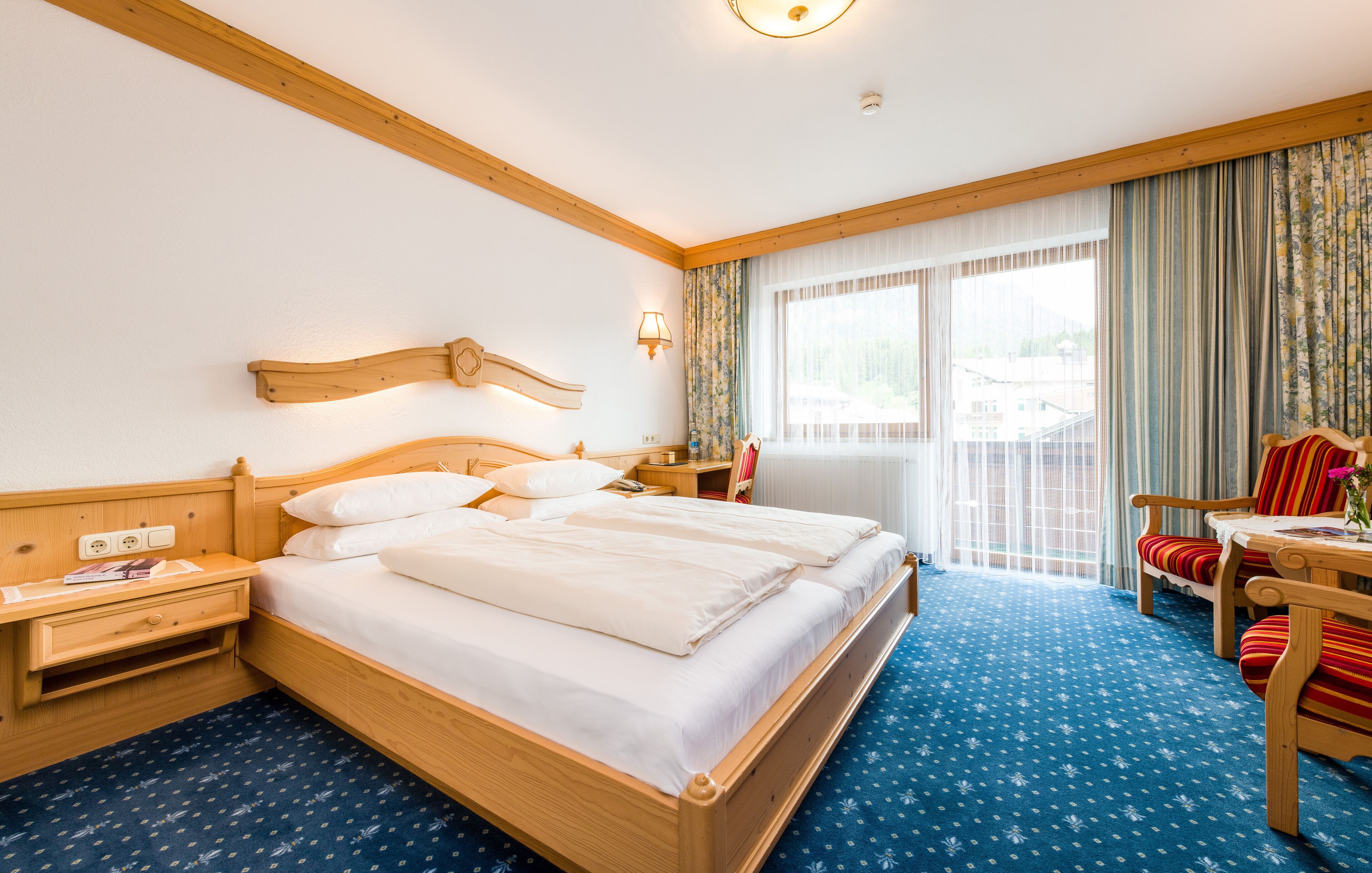 Ein Hotelzimmer mit einem hölzernen Bett, einem Nachttisch, einem Schreibtisch mit Stuhl und einem Fenster mit Vorhängen.