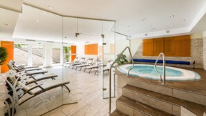 Un espace piscine intérieur moderne, doté de parois en verre transparent, de chaises longues et d’un mur en pierre agrémenté de panneaux orange.