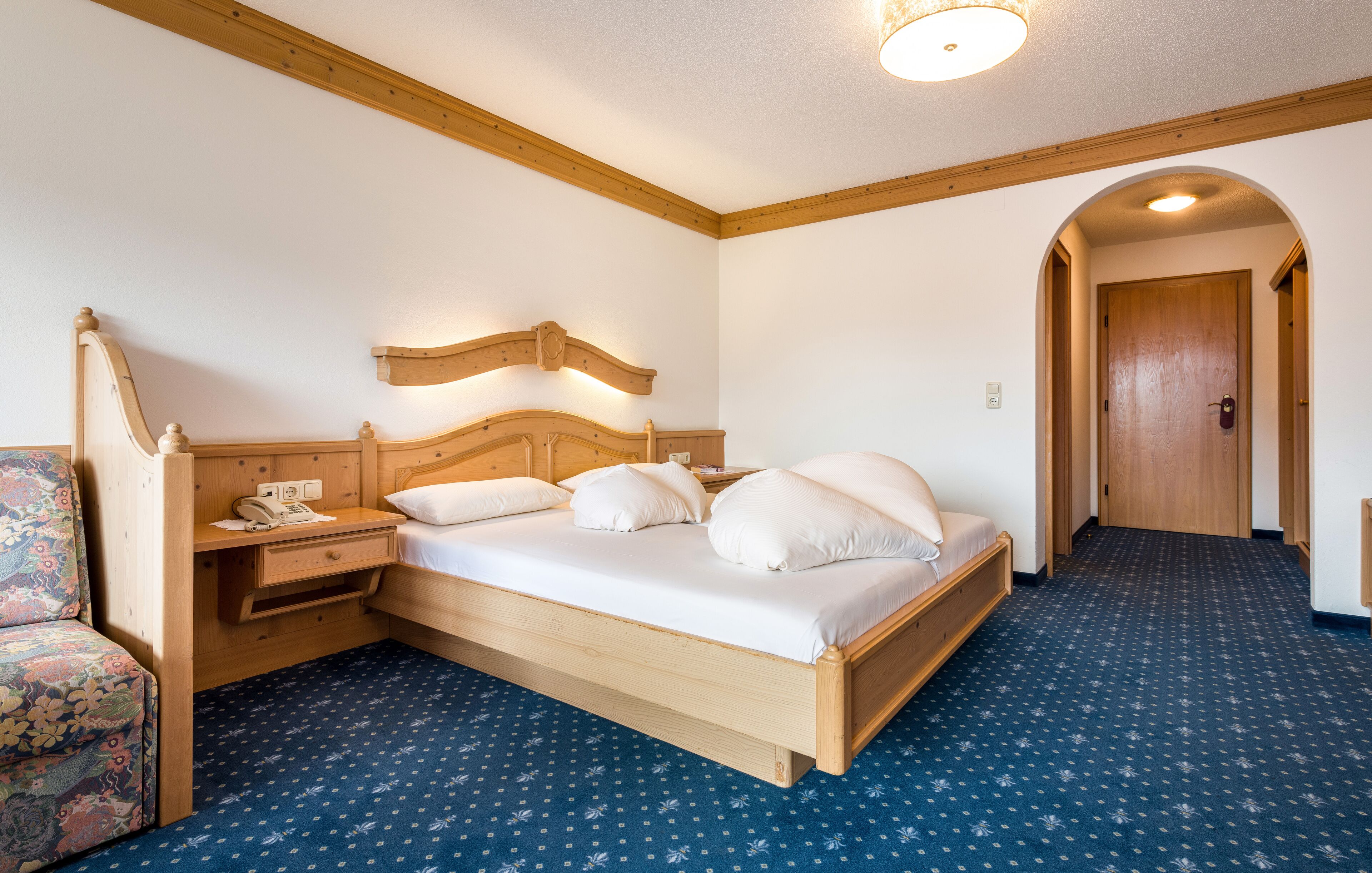 A bedroom with a wooden bed, a nightstand, a telephone, and a floral chair.