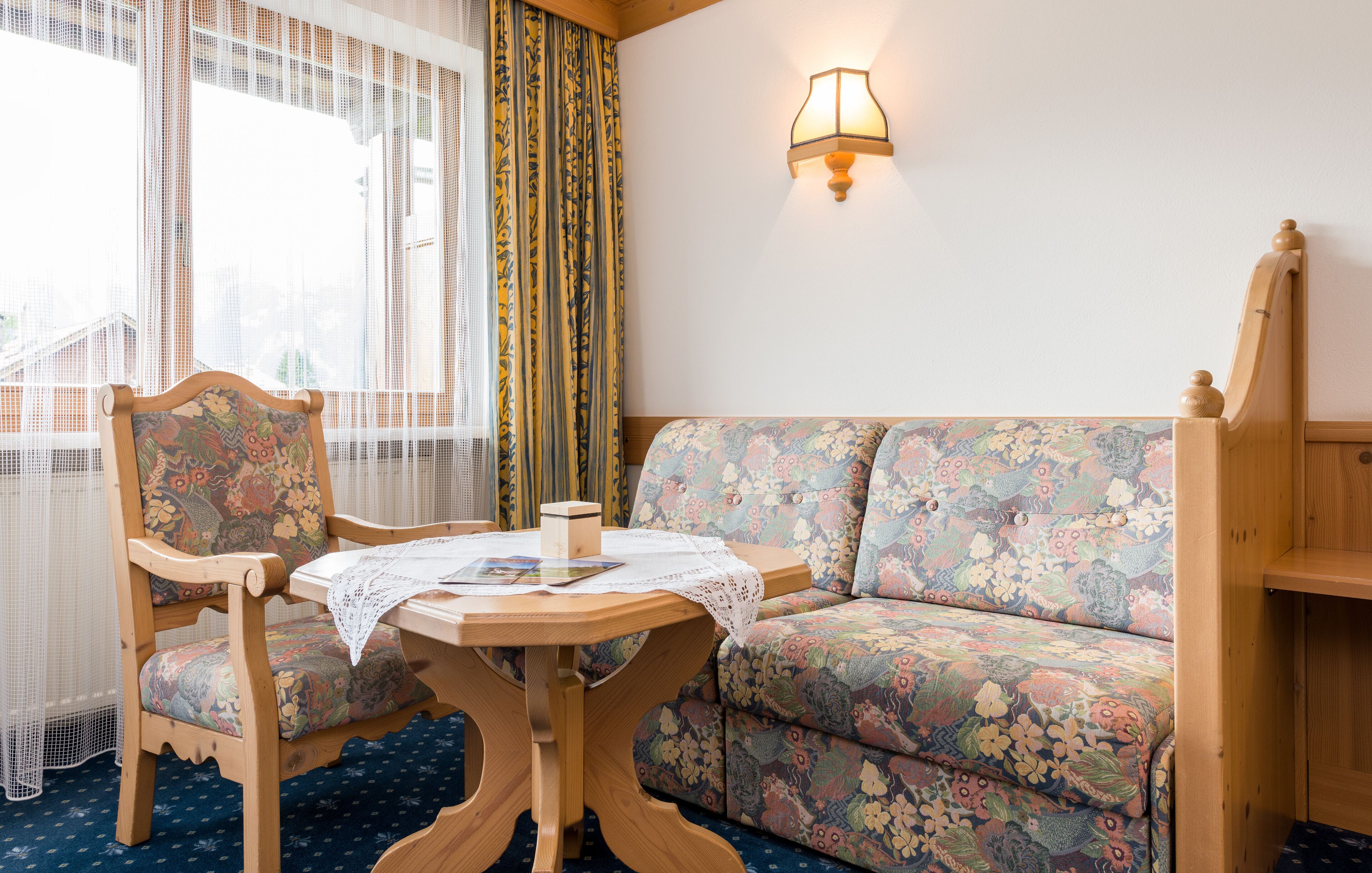 A room with a floral sofa, a wooden chair, a small table with a lace tablecloth, and a wall-mounted lamp.