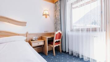 A hotel room with a bed, a desk with a telephone, a chair, and a window with sheer curtains.