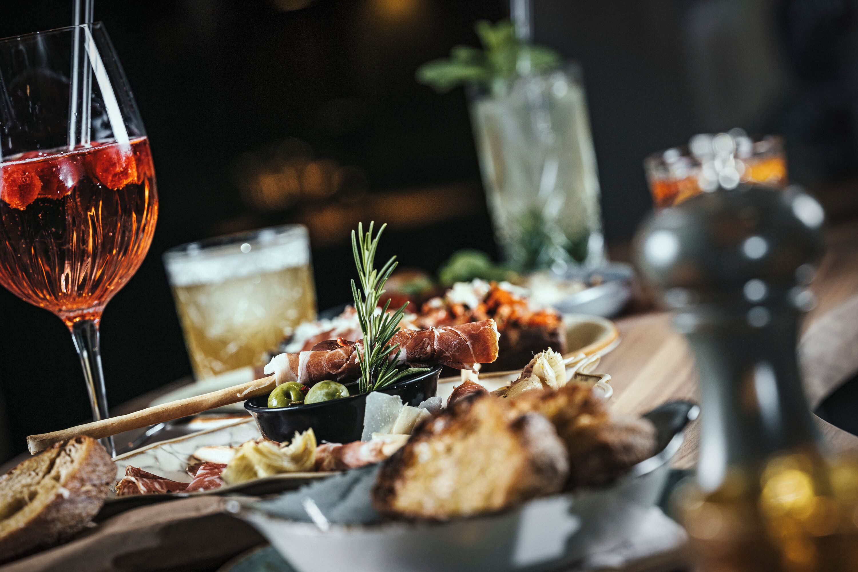 Una mesa con diversos alimentos, entre ellos pan, queso y embutidos, acompañada de copas de vino.