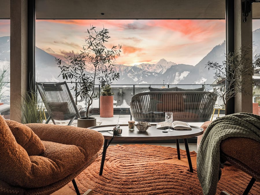 Una sala de estar moderna con un gran ventanal con vistas a montañas, que incluye una mesa redonda, dos sillas y una planta en maceta.