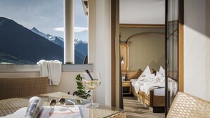 A balcony with a view of mountains, a wicker table with a glass of wine, and a bed in the background. - Hotel Sieghard (Schwendau)