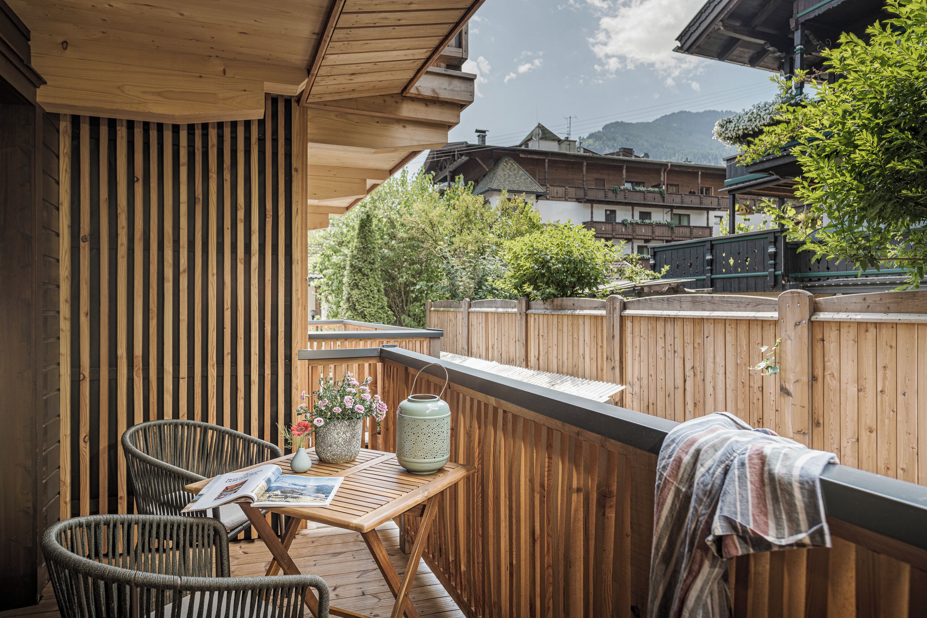 Une terrasse en bois avec des chaises en osier, une table avec un magazine, une lanterne et une serviette posée sur la rambarde.