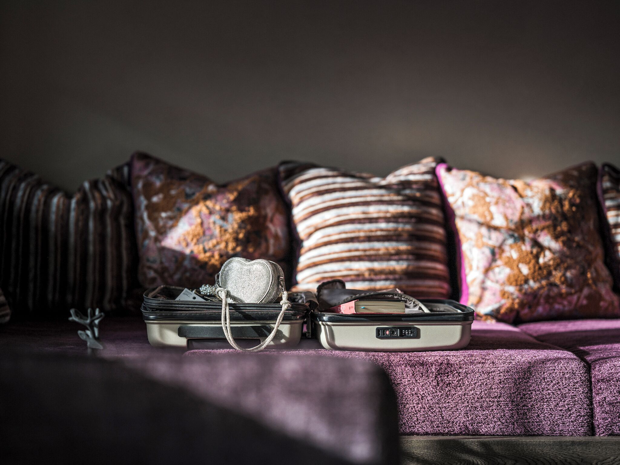 Una cama con una manta morada y almohadas, un pequeño bolso plateado con forma de corazón, y una maleta negra sobre la cama.