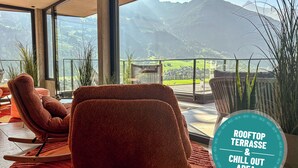 A rooftop terrace with a view of mountains, furnished with a rocking chair and a cushioned chair, and a potted plant. - Hotel Sieghard (Schwendau)
