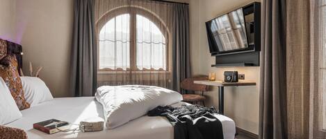 Una habitación de hotel con una cama grande, un televisor montado en la pared, una mesita con una lámpara y una ventana con cortinas.