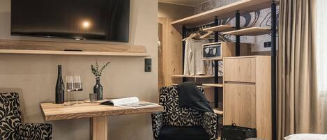 Habitación de hotel moderna con televisor de pantalla plana, escritorio de madera, dos sillas estampadas, cama con manta gris y estanterías empotradas con ropa y una radio.