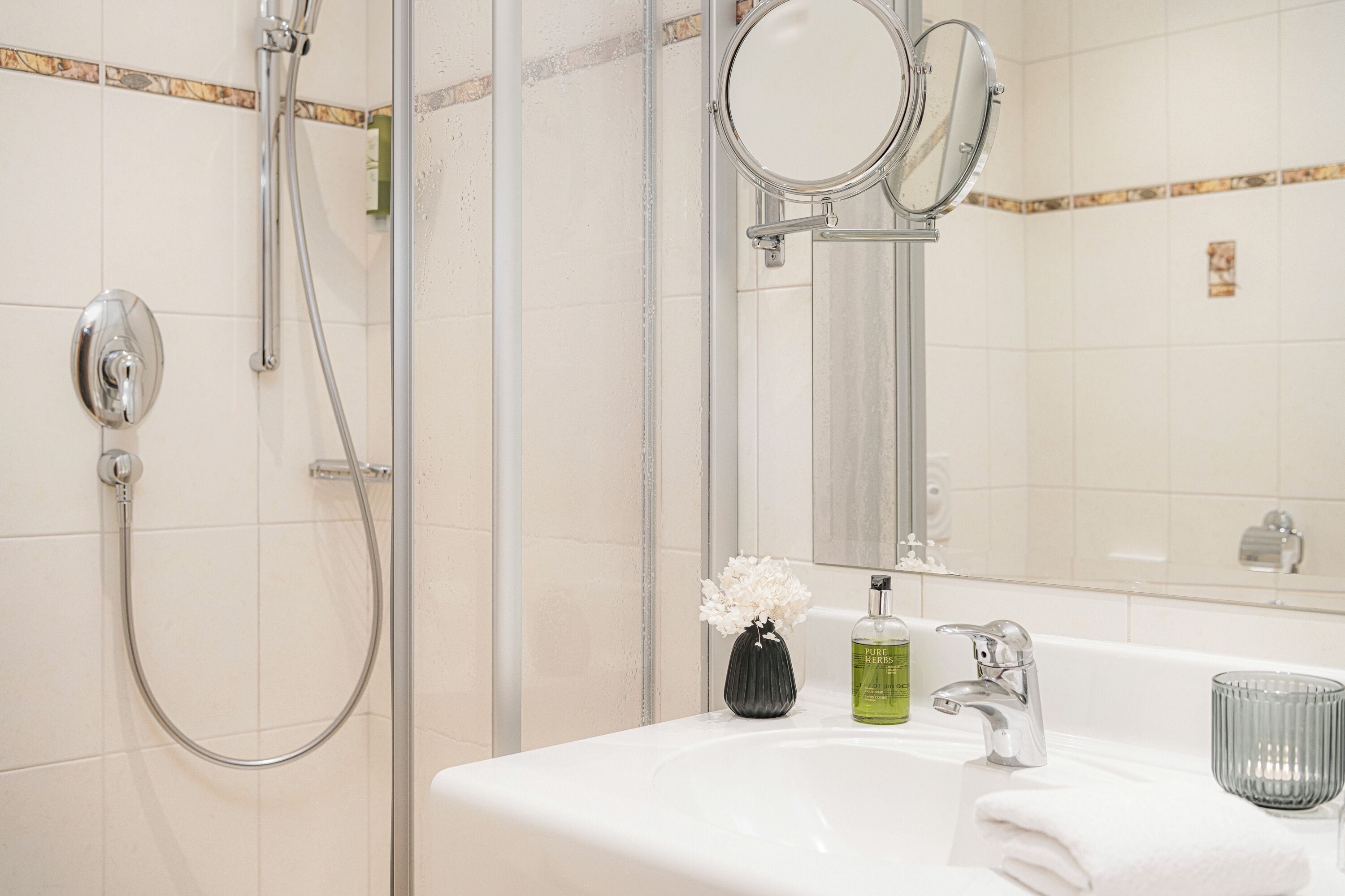 Un baño con ducha, lavamanos, espejo y un florero con flores.
