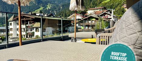 A rooftop terrace with a view of a mountain village, outdoor seating, and a sign indicating the area.