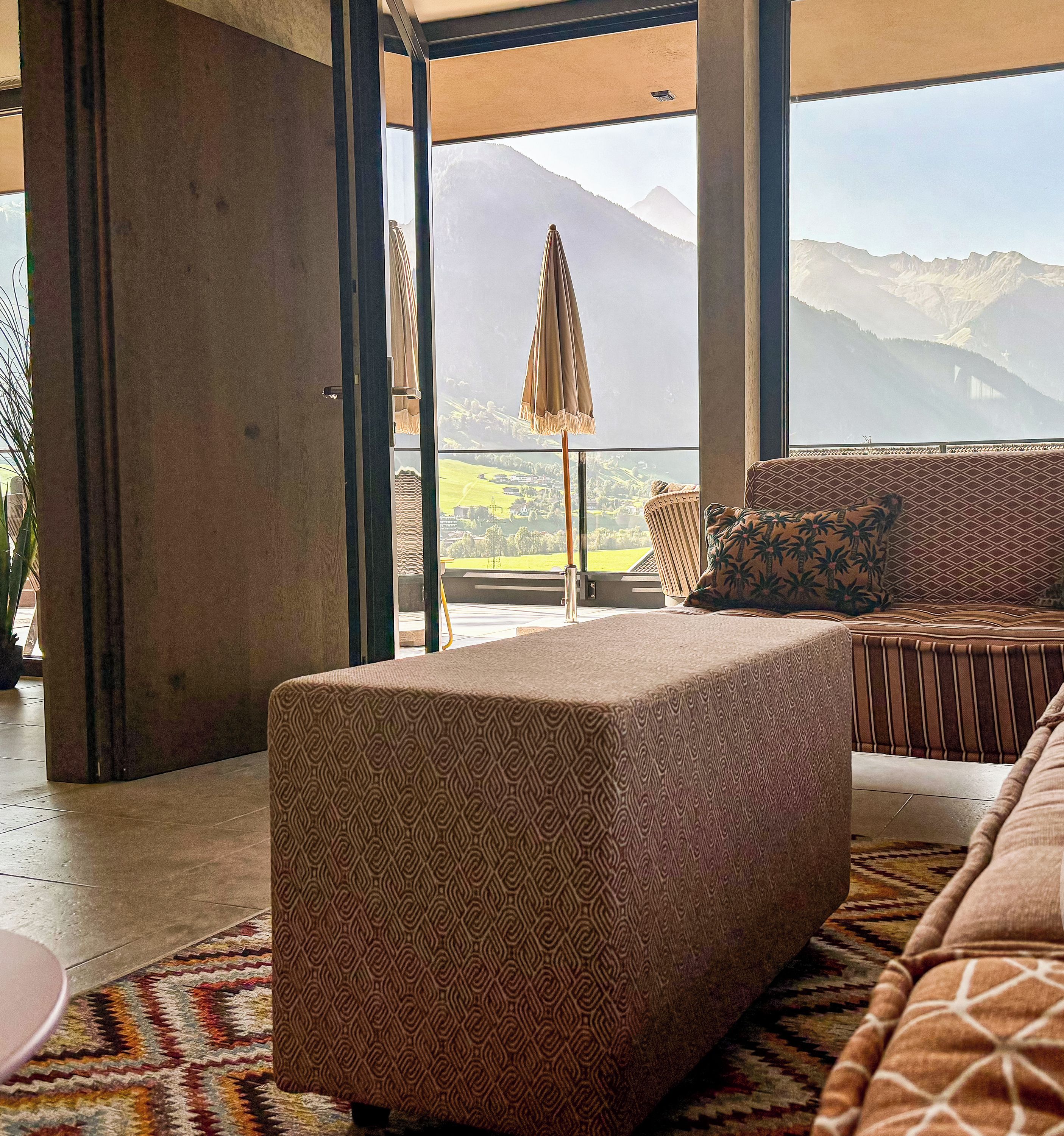 A modern living room with a large window overlooking a mountainous landscape, a patterned sofa, and a small table.